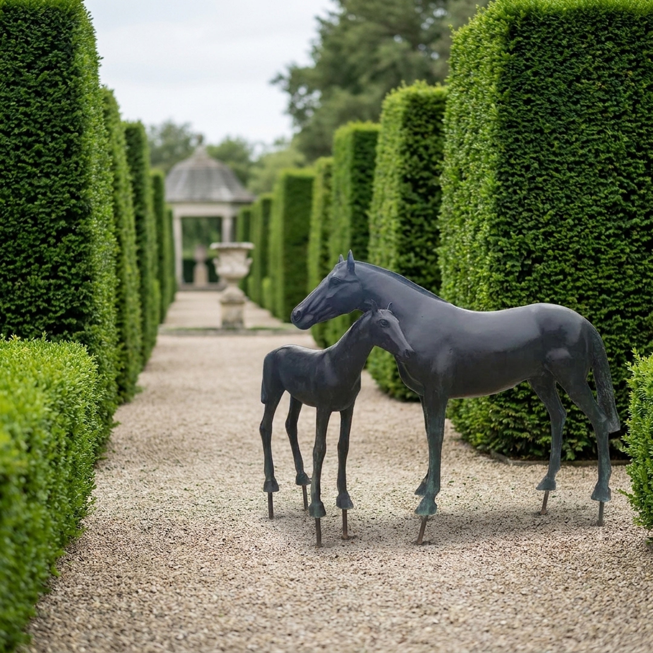 Life-Size Mare and Foal Bronze Sculpture by Giant Sculptures with dark patina, displayed on a gravel path between tall topiary hedges leading to a garden pavilion.