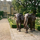 Life-Size Pair of African Elephants Bronze Sculpture by Giant Sculptures with warm reddish-brown patina, displayed on a stone path beside a rose garden and manor house.
