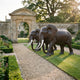 Life-Size Pair of African Elephants Bronze Sculpture by Giant Sculptures, dark patina with ivory-white tusks, displayed on lawn beside a gravel path and stone archway.