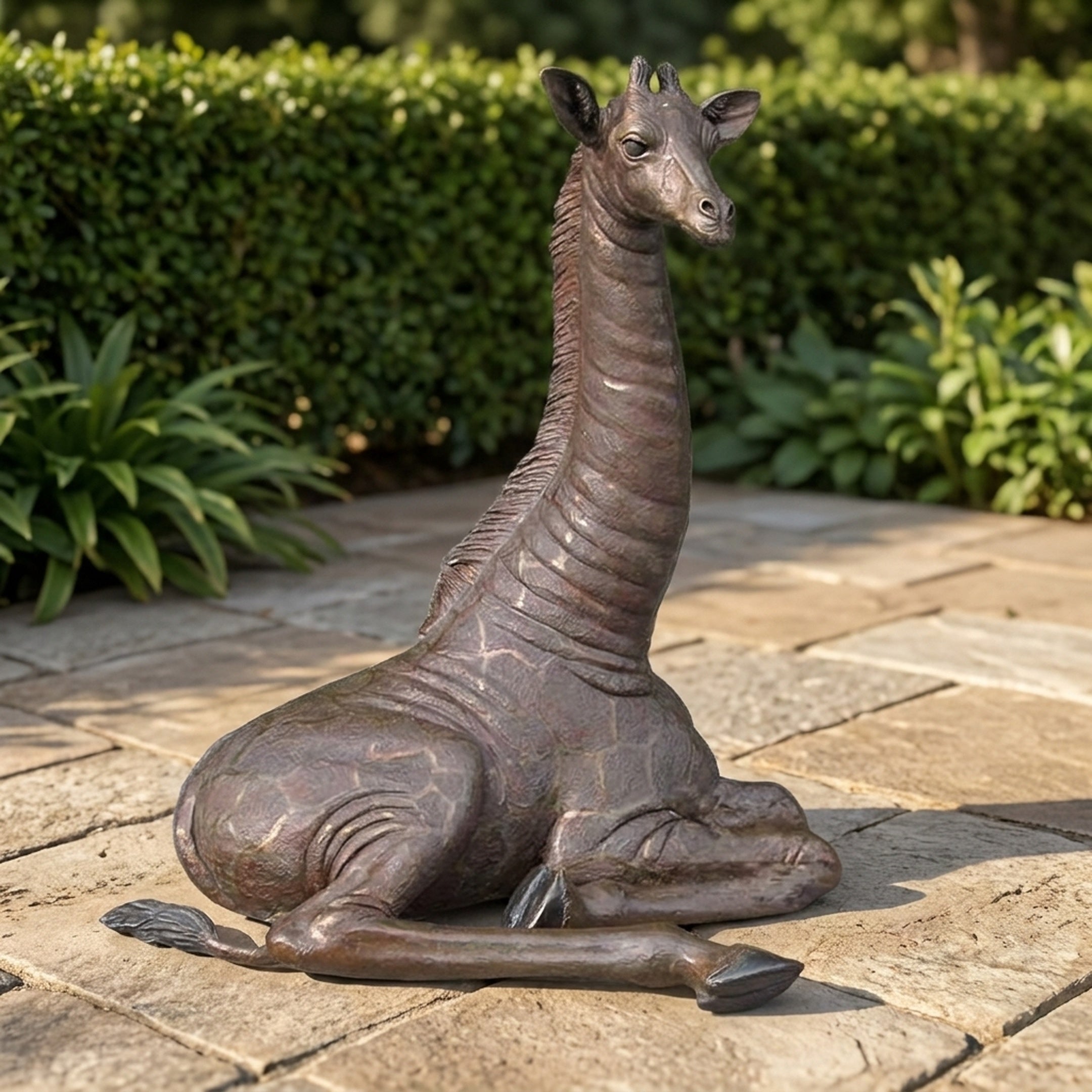 Life-Size Resting Baby Giraffe Bronze Sculpture by Giant Sculptures, 120cm, in dark brown patina, shown in a folded-leg resting pose on a stone patio beside hedging.