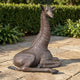 Life-Size Resting Baby Giraffe Bronze Sculpture by Giant Sculptures, 120cm, in dark brown patina, shown in a folded-leg resting pose on a stone patio beside hedging.
