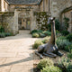 Life-Size Resting Baby Giraffe Bronze Sculpture by Giant Sculptures in dark brown patina, folded-leg pose on gravel beside a stone pathway and manor entrance.