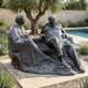Life-Size Seated Conversing Figures Bronze Sculpture by Giant Sculptures, 220cm, shown in a sunlit garden with lavender, olive tree, and pool behind.