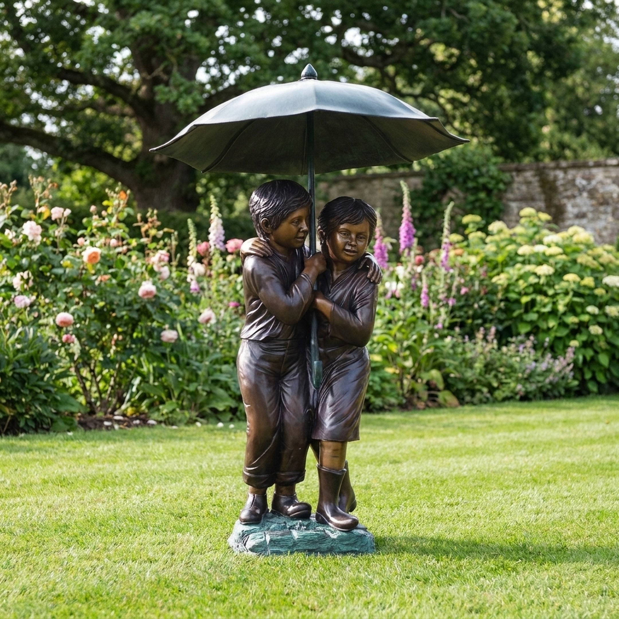 Life-Size Traditional Boy and Girl Bronze Sculpture by Giant Sculptures, 190cm, two children sheltering under an umbrella on a garden lawn beside flowerbeds.
