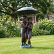 Life-Size Traditional Boy and Girl Bronze Sculpture by Giant Sculptures, 190cm, two children sheltering under an umbrella on a garden lawn beside flowerbeds.