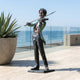 Life-Size Traditional Boy Violinist Bronze Statue by Giant Sculptures with dark patina and green verdigris jacket, displayed on a seaside terrace beside an agave planter.