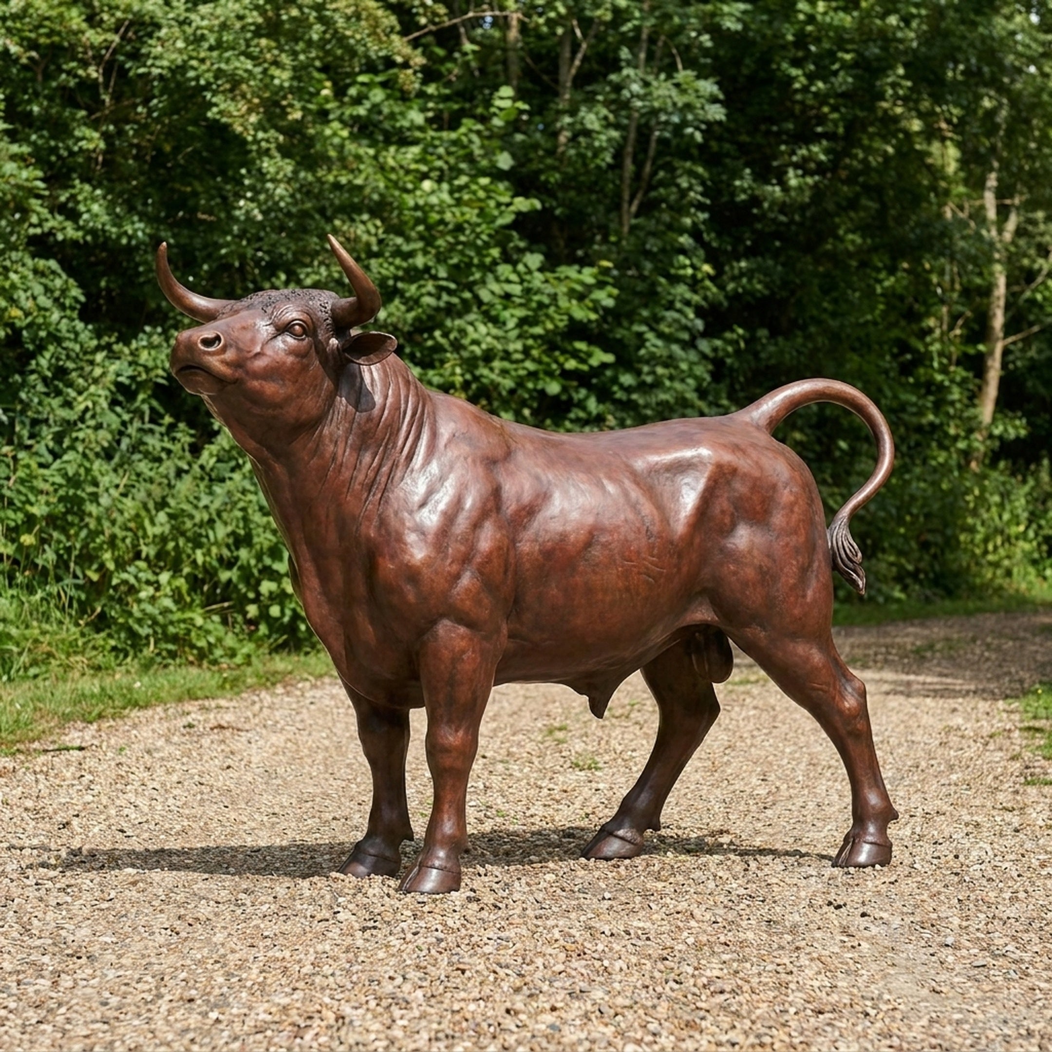 Life-Size Traditional Bull Bronze Sculpture by Giant Sculptures shown side-on with raised head and curled tail, reddish-brown patina, displayed on gravel near trees.