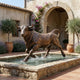 Life-Size Traditional Charging Bull Bronze Sculpture by Giant Sculptures, warm brown patina, mid-stride in a stone fountain basin in a Mediterranean courtyard.