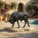 Life-Size Traditional Charging Bull Bronze Sculpture by Giant Sculptures, head lowered in mid-charge on a stone slab base beside a poolside Mediterranean courtyard.