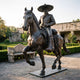 Life-Size Traditional Charro Rider and Horse Bronze Sculpture by Giant Sculptures in dark patina, prancing pose, displayed on a stone courtyard near a carved bench.