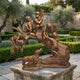Life-Size Traditional Children Playing on a Tree Log Bronze Sculpture by Giant Sculptures, displayed on a stone plinth in a Mediterranean garden with olive trees.