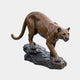 Life-Size Traditional Crouching Cougar Bronze Sculpture by Giant Sculptures, golden-brown patina cat descending a dark stone base, shown against plain grey.