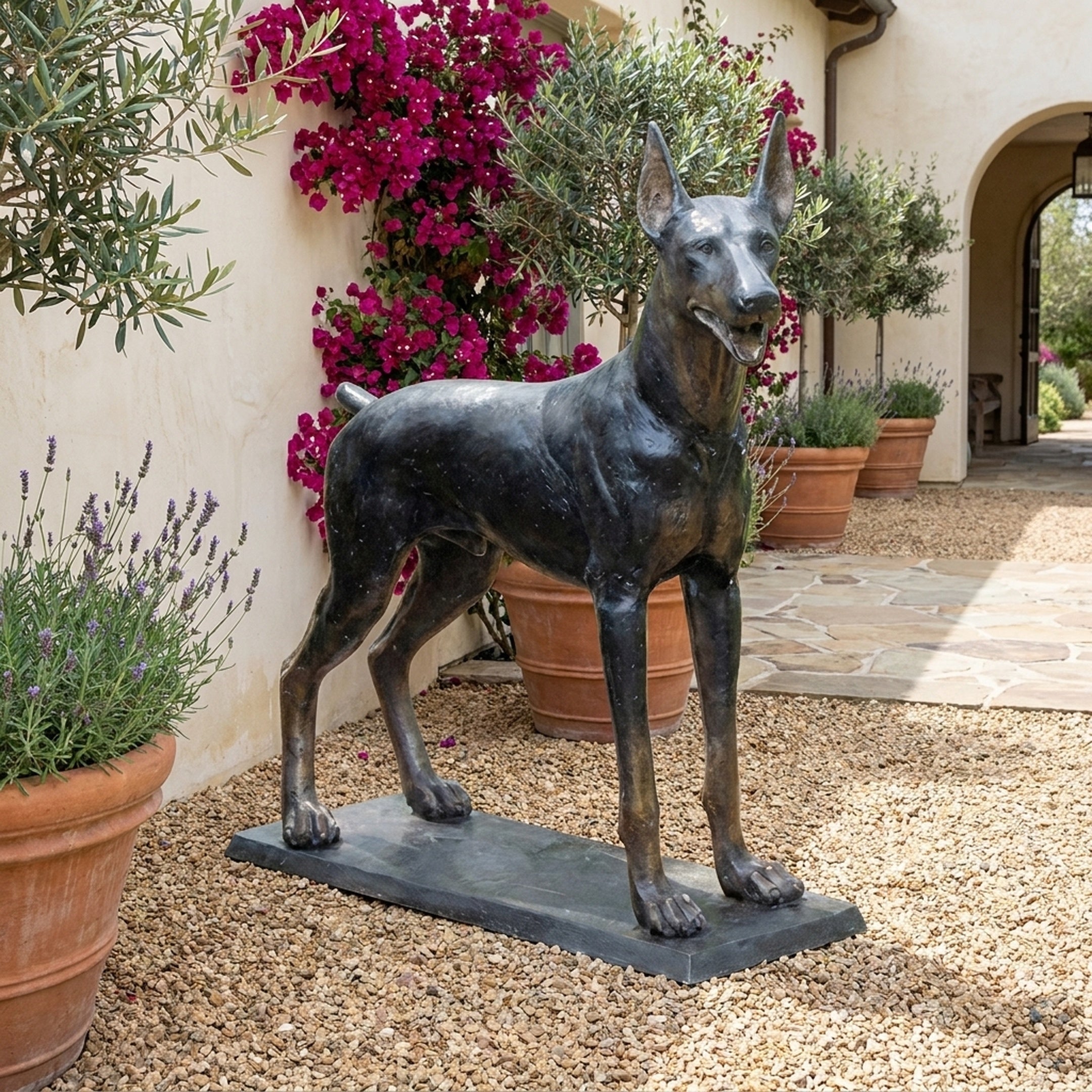 Life-Size Traditional Doberman Pinscher Bronze Sculpture by Giant Sculptures on a flat base, displayed on gravel beside lavender, bougainvillea, and terracotta pots.