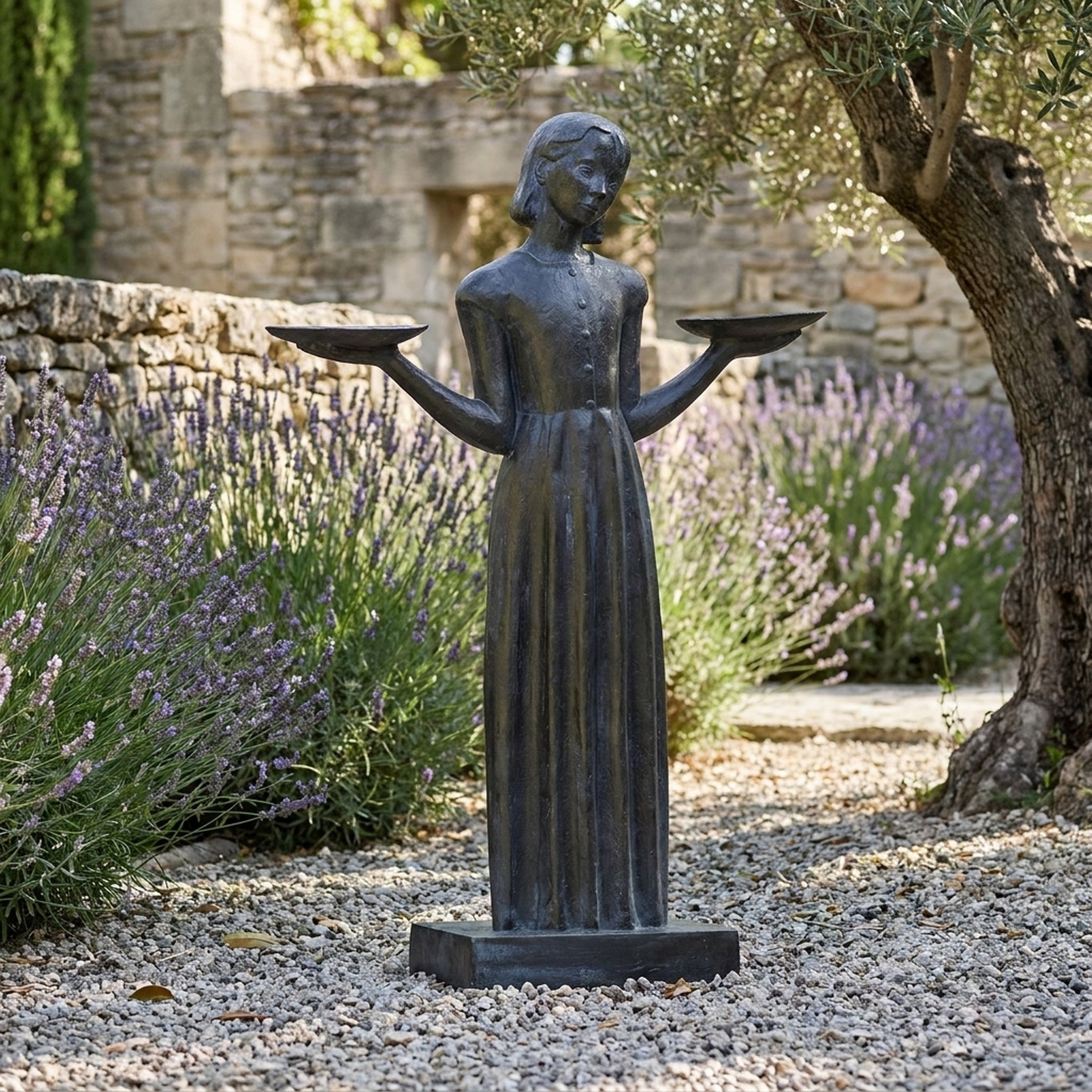 Life-Size Traditional Girl with Bird Trays Bronze Sculpture by Giant Sculptures, set on gravel beside lavender and an olive tree in a stone-walled garden.