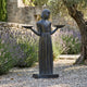Life-Size Traditional Girl with Bird Trays Bronze Sculpture by Giant Sculptures, set on gravel beside lavender and an olive tree in a stone-walled garden.