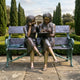 Life-Size Traditional Girls on Bench Bronze Sculpture by Giant Sculptures, showing two barefoot girls writing in a book together on an iron-framed bench in a formal garden.