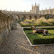 Life-Size Traditional Greyhound Bronze Sculpture by Giant Sculptures resting on a lawn in a historic college garden courtyard with stone arches behind.