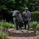 Life-Size Traditional Highland Bull and Herdsman Bronze Sculpture by Giant Sculptures, dark patina, set on stones in a garden bed with foxgloves and hedging.