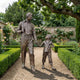 Life-Size Traditional Man and Boy Fishing Bronze Sculpture by Giant Sculptures on a gravel garden path, with rose beds, clipped hedges, and an overhanging tree.