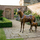 Life-Size Traditional Mare and Foal Bronze Sculpture by Giant Sculptures, warm brown patina, displayed on a stone courtyard path beside roses and a brick wall.