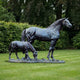 Life-Size Traditional Mare and Foal Bronze Sculpture by Giant Sculptures with blackened patina, mare standing alert beside grazing foal on a lawn before green hedging.