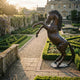 Life-Size Traditional Rearing Horse Bronze Sculpture by Giant Sculptures with reddish-brown patina, displayed in a formal manor garden with clipped hedging at sunset.