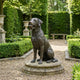 Life-Size Traditional Sitting Labrador Bronze Sculpture by Giant Sculptures holding a ball, displayed on a round stone base in a hedged gravel garden path.
