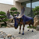 Life-Size Standing Bull Moose Bronze Sculpture by Giant Sculptures with iridescent purple and amber patina, displayed on gravel beside bonsai pines and stones.