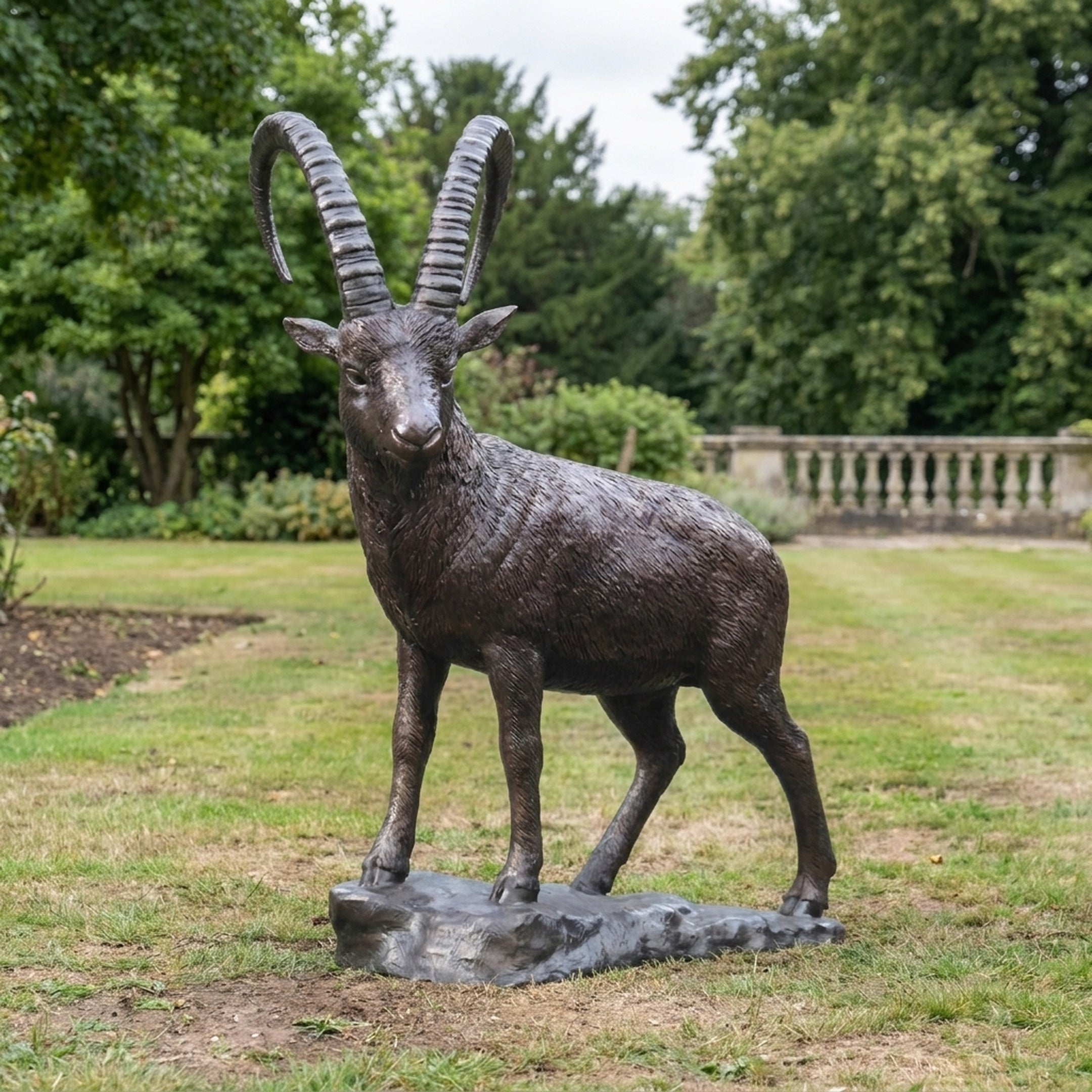 Life-Size Traditional Standing Ibex Bronze Sculpture by Giant Sculptures with tall ridged horns and dark patina, displayed on grass in a formal garden.