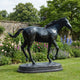 Life-Size Traditional Trotting Horse Bronze Sculpture by Giant Sculptures, dark patina, shown trotting on an oval base in a garden beside flower beds.