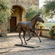 Life-Size Traditional Trotting Horse Bronze Sculpture by Giant Sculptures, shown mid-stride with dark brown patina on a stone plinth in a Tuscan courtyard.