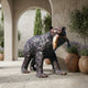 Life-Size Traditional Walking Bear Bronze Sculpture by Giant Sculptures, holding a salmon in its jaws, displayed on a stone courtyard beside potted olive trees and lavender.