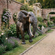 Life-Size Walking African Elephant Bronze Sculpture by Giant Sculptures, shown mid-stride in a walled garden beside lavender, roses, and a gravel path.