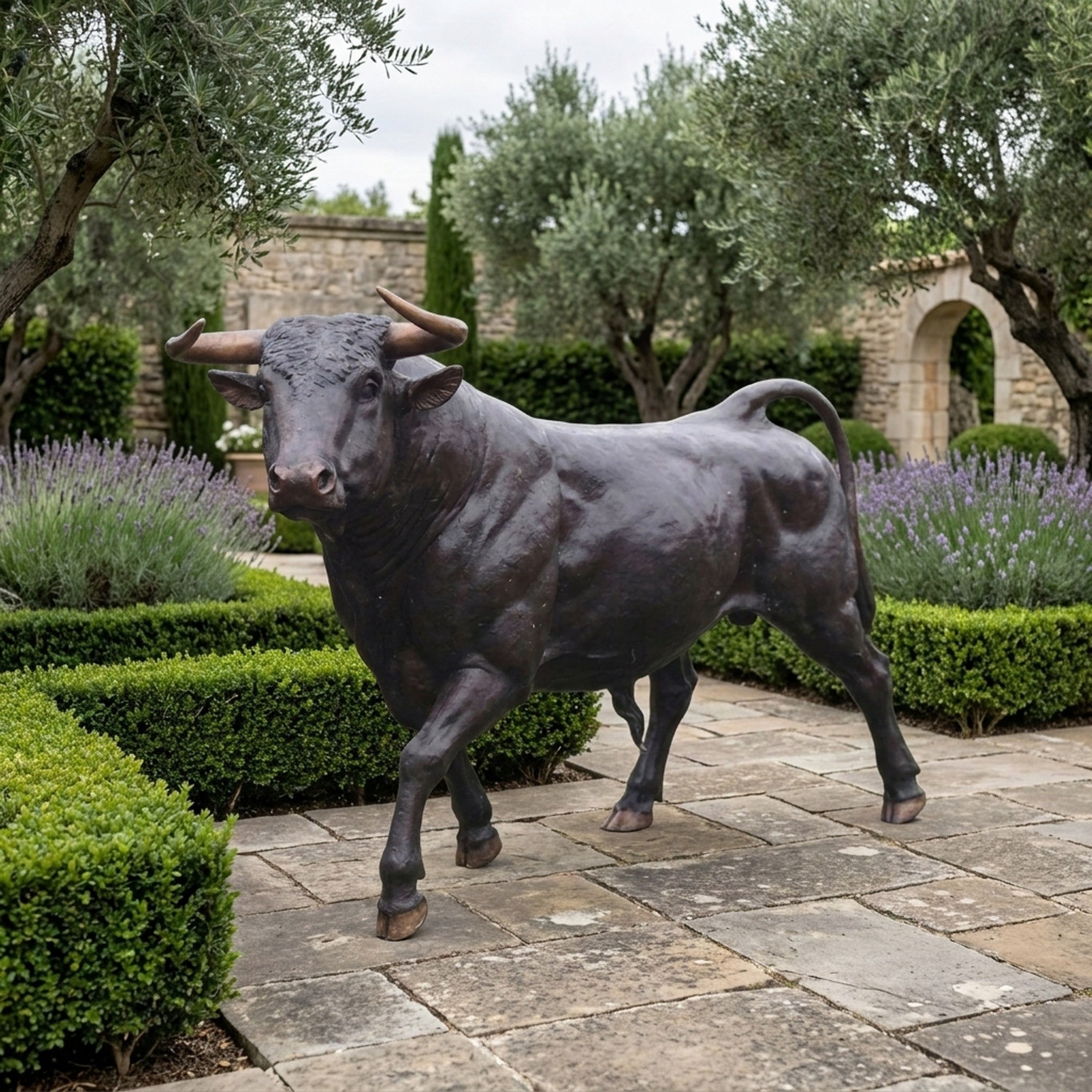Life-Size Walking Longhorn Bull Bronze Sculpture by Giant Sculptures with dark patina, displayed on a stone courtyard among lavender, boxwood, and olive trees.