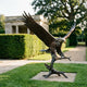 Monumental Bald Eagle Bronze Sculpture by Giant Sculptures, 280cm, wings raised mid-landing on a branch perch, displayed on a lawn beside a manicured hedge.