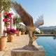 Giant Sculptures Monumental Bald Eagle Landing Bronze Sculpture with wings raised on a stone plinth beside a pool, with bougainvillea and hillside views.