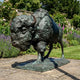 Monumental Charging Bison Bronze Sculpture by Giant Sculptures, side view with lowered head and verdigris patina, set on a stone patio beside a flower garden.