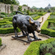 Monumental Charging Bull Bronze Sculpture by Giant Sculptures with black patina and gold horns, displayed on a wooden base in a formal hedged garden.