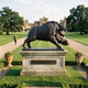 Monumental Charging Wild Boar Bronze Sculpture by Giant Sculptures on a stone plinth along a grand estate lawn, with a man walking the gravel path behind.