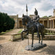 Monumental Classical Armoured Knight on Horseback Bronze Sculpture by Giant Sculptures, dark patina, lance raised, prancing horse, displayed in a colonnaded courtyard.