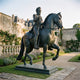 Monumental Classical Armoured Rider and Rearing Horse Bronze Sculpture by Giant Sculptures, dark patina, displayed in a château garden with roses.