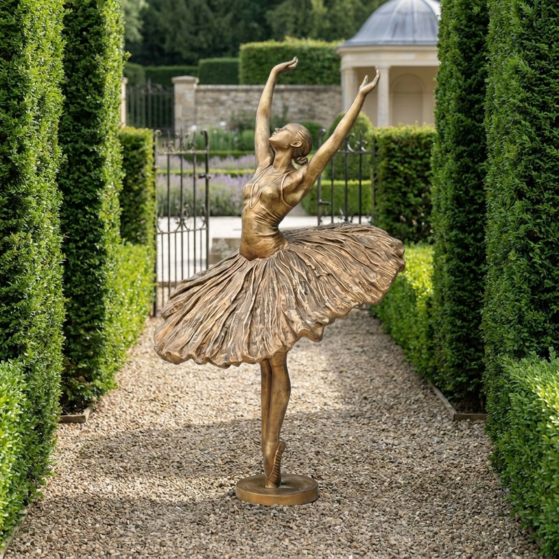 Monumental Classical Ballerina Dancer Bronze Sculpture by Giant Sculptures in golden-bronze finish, posed en pointe with arms raised, on a gravel garden path.