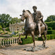 Monumental Classical Bearded Rider and Horse Bronze Sculpture by Giant Sculptures, 430cm, shown in a formal garden with rose beds and a stone pavilion behind.