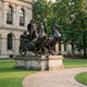Monumental Classical Chariot with Goddess and Horses Bronze Sculpture by Giant Sculptures, shown on a stone plinth outside a classical building with trees behind.