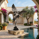 Monumental Classical Cupid Archer Bronze Sculpture by Giant Sculptures, dark patinated winged figure drawing a bow, on a stone plinth beside a Mediterranean courtyard pool.
