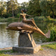 Monumental Classical Female Figure on Dolphin Bronze Sculpture by Giant Sculptures, with gold-accented ruffles, on a rough stone plinth beside a lake.