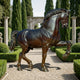 Monumental Classical Horse Bronze Sculpture by Giant Sculptures, dark-patinated stallion mid-prance on a gravel garden path lined with cypress trees and hedges.