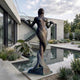 Monumental Classical Mermaid Bronze Sculpture by Giant Sculptures with verdigris patina, displayed beside a reflecting pool in a modern concrete courtyard.