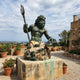 Monumental Classical Poseidon Bronze Sculpture by Giant Sculptures with verdigris patina, trident raised and sea turtle in hand, on a rocky base in a coastal garden.