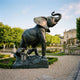 Monumental Classical Rearing Elephant Bronze Sculpture by Giant Sculptures, trunk raised with a lion at its base, displayed on gravel beside a chateau garden pond.